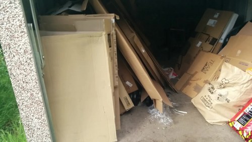 A partially enclosed outdoor space, likely a garage or storage area, containing various discarded items including a boxy, beige piece of furniture or panel leaning against the left wall, with a rough textured concrete floor. To the right, there are multiple large cardboard boxes stacked haphazardly, some with visible labels and printed text, and a paper bag filled with miscellaneous waste. Scattered plastic wrapping and debris are also visible on the ground. The area appears cluttered and in typical condition for waste awaiting collection or removal. The background is dark, indicating limited lighting, and the environment suggests a residential or commercial premises in the Palmers Green area. Business Waste Removal Palmers Green can assist with clearing such rubbish from properties within N13 postcode, supporting local waste management needs.