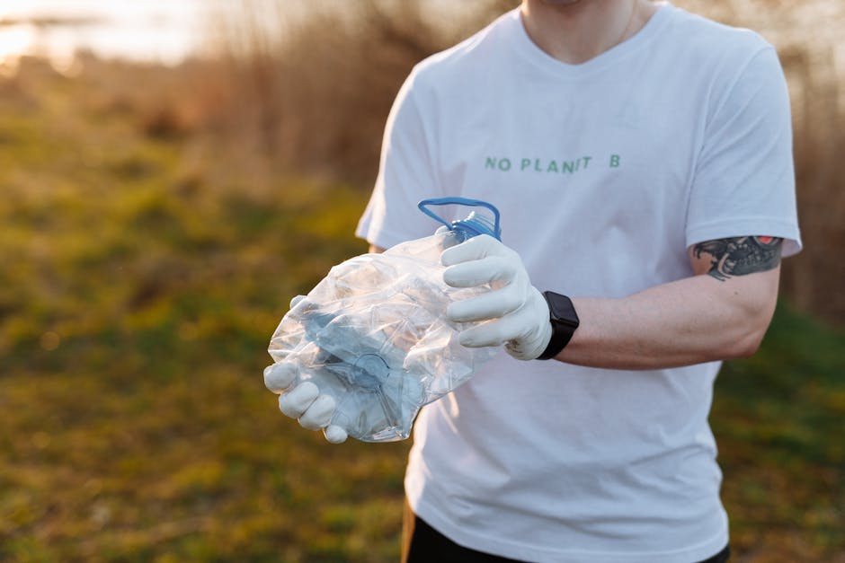 A person wearing a white T-shirt with the words 'NO PLANT B' printed on it and a black wristwatch is standing outdoors on a grassy area during daylight. The individual is holding a crumpled transparen